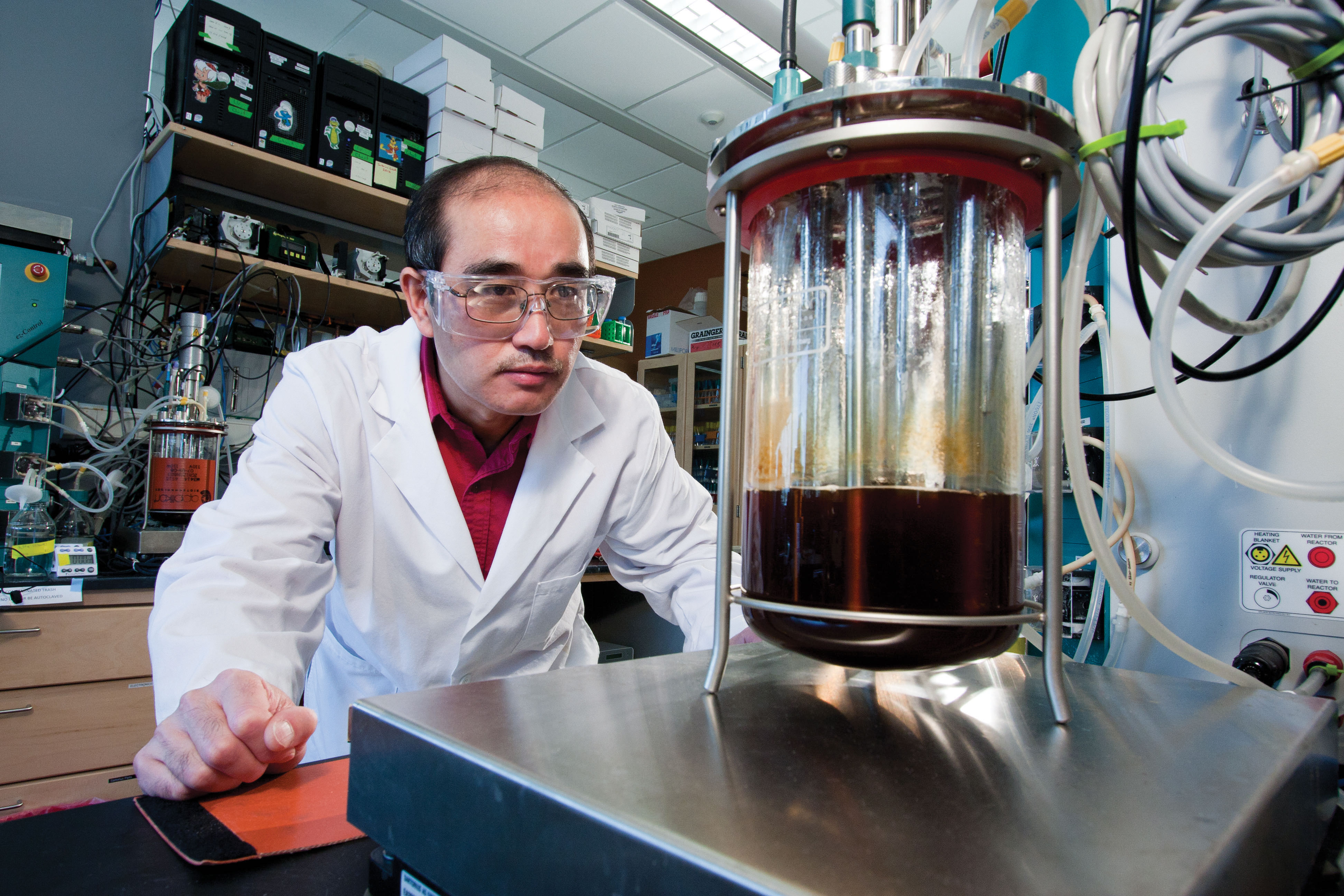 Man in white lab coat and protective glasses peers at flask of brown liquid in lab