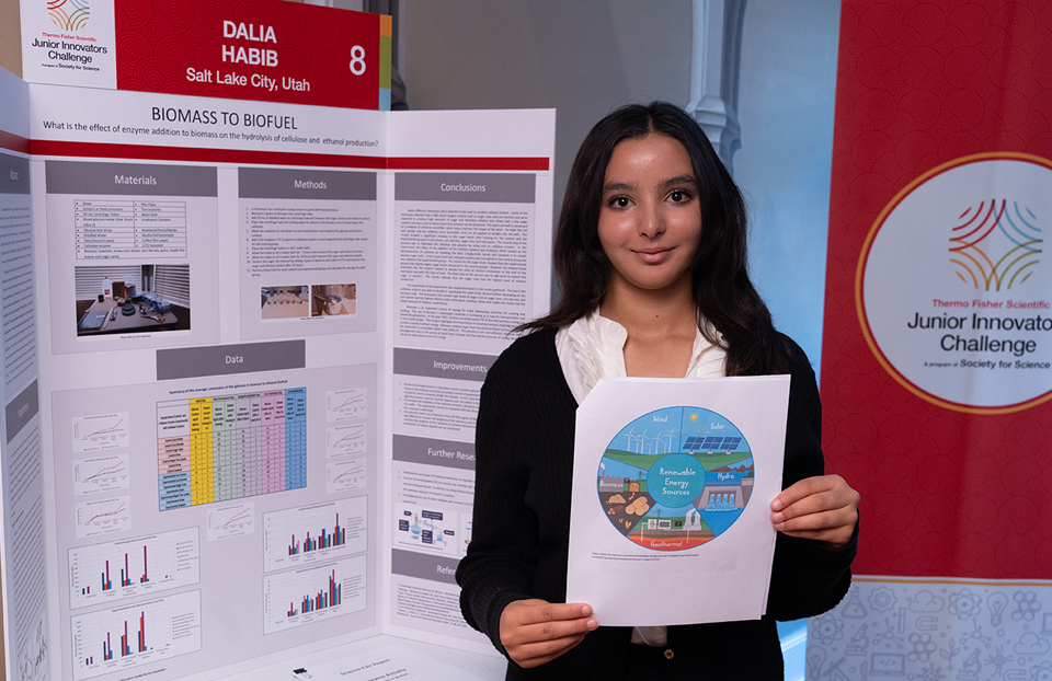 Girl standing in front of a scientific poster holding a sheet of paper showing various renewable energy sources