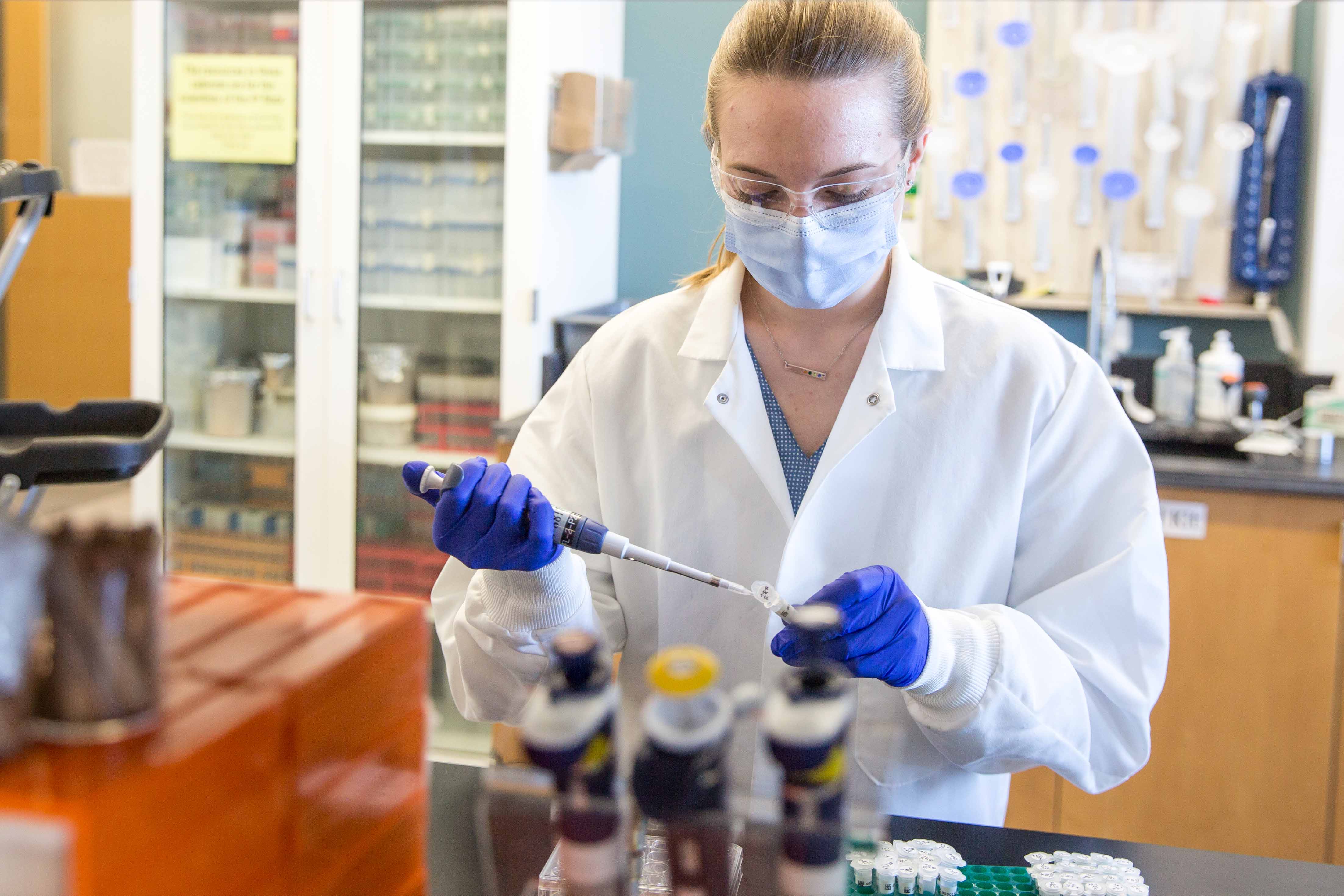 Amy Enright pipettes in a lab coat, gloves, and facemask.