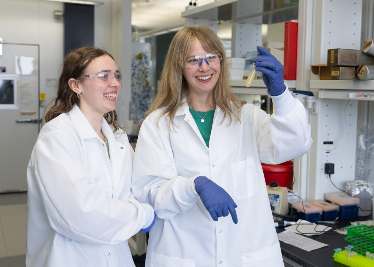 A woman in a white lab coat and safety glasses holds up a flask of opaque yellow liquid as another woman in similar attire looks on smiling