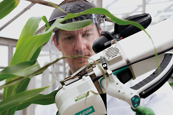 Man with a clear face visor looks at a piece of equipment. His face is framed by the instrument and green plant leaves
