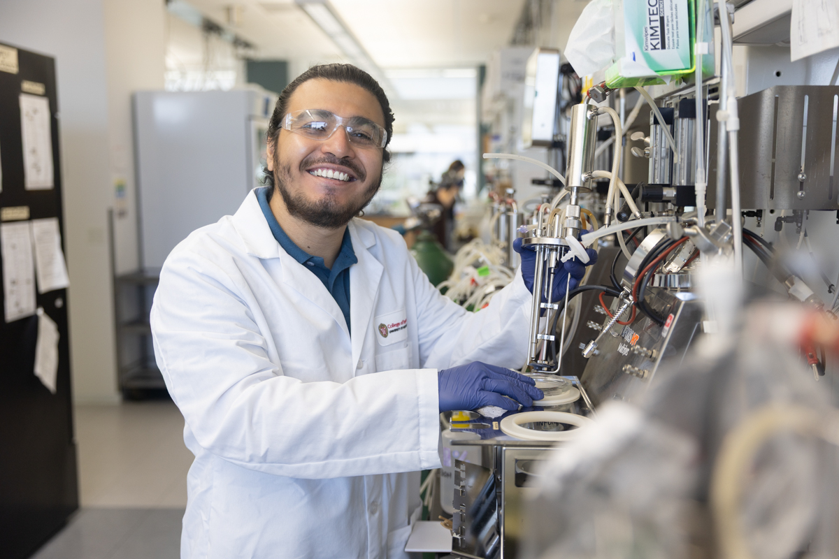 Smiling man in white lab coat and purple nitrile gloves holds a piece of equipment in a modern lab