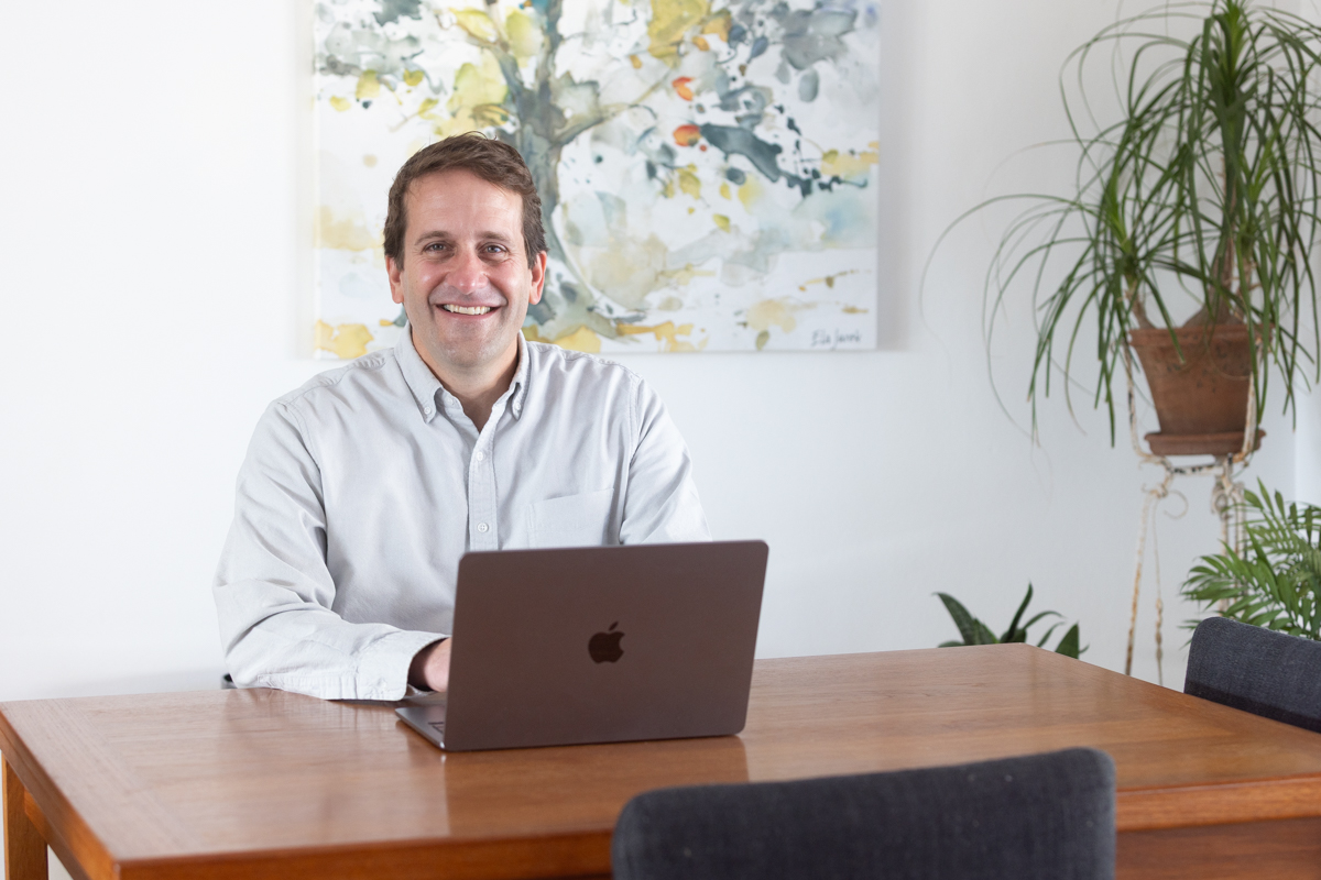 A smiling man sitting at a desk behind an open laptop computer