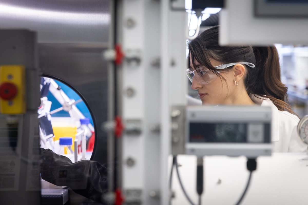 A researcher in a white lab coat and safety goggles looks through a circular portal into an automated laboratory workstation filled with robotic equipment and chemical vials.