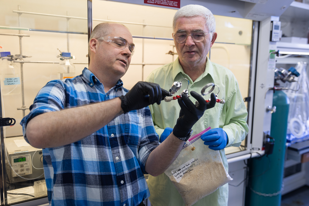 Man wearing black rubber gloves holding a piece of lab equipment as man holding plastic bag of wood chips looks on