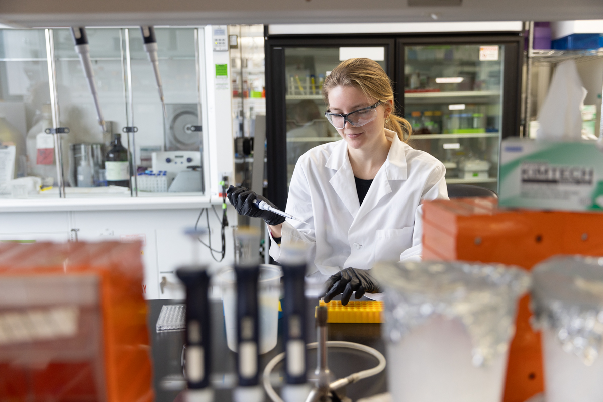 A woman in a white lab coat and lab glasses holds a pipette at a lab bench