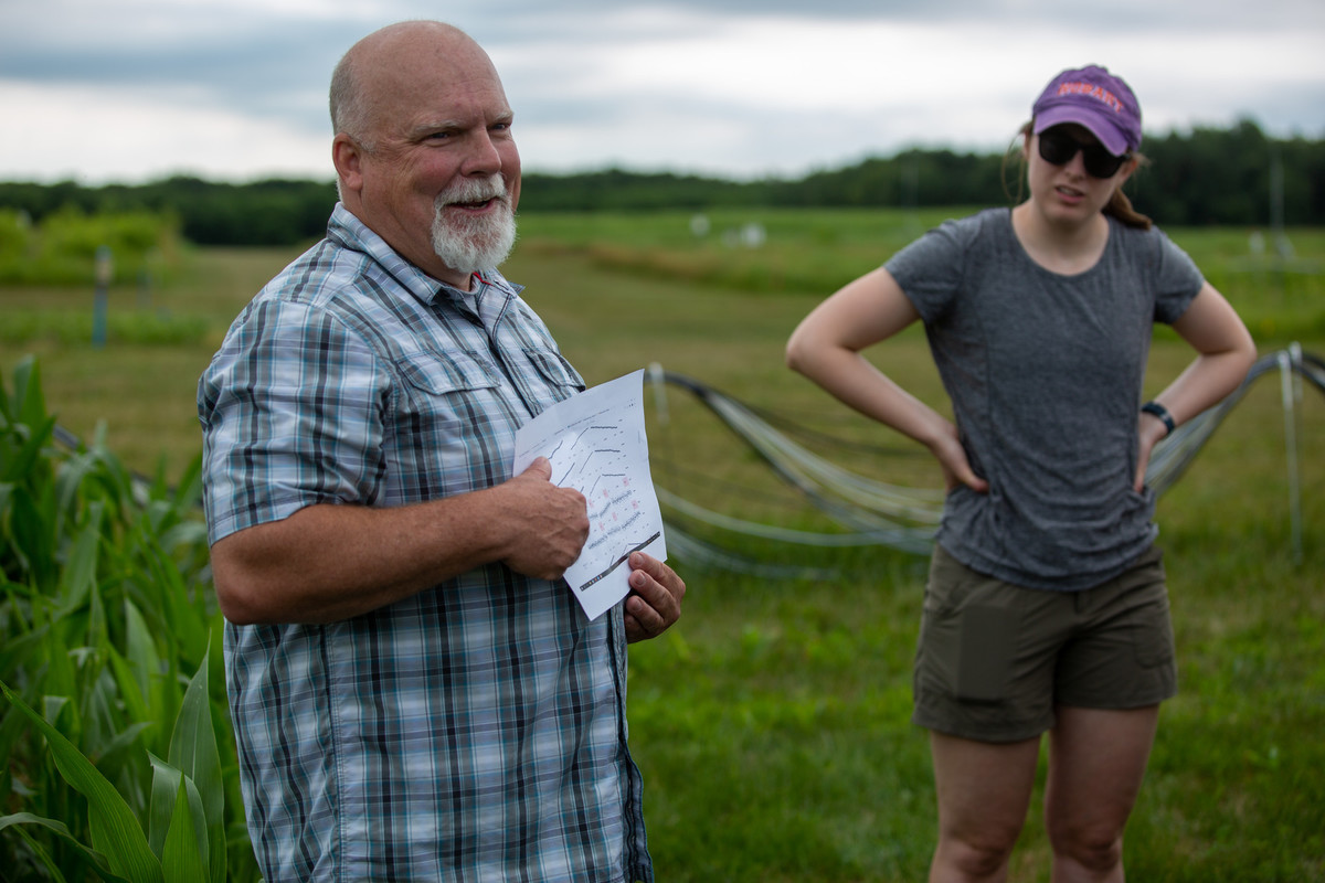Man with white goatee in blue checked shirt stands in green field pointing to text on a piece of paper