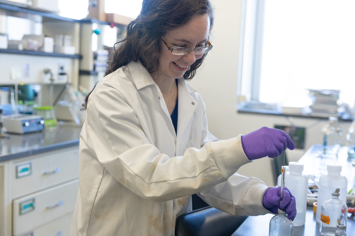 Woman in white lab coat and purple latex gloves opens a glass container on a black counter in a