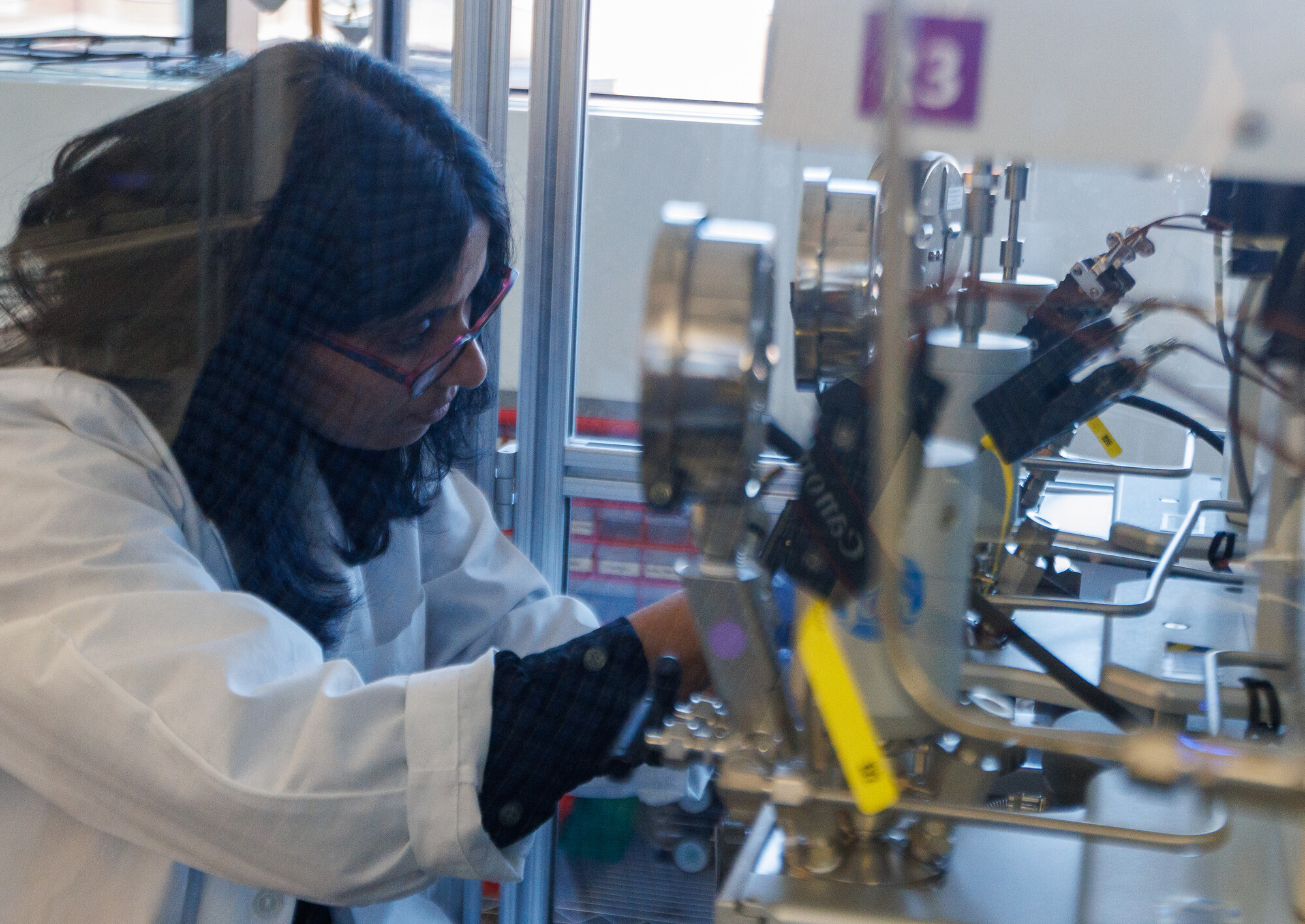 A researcher in a lab coat, safety glasses, and nitrile gloves sets up a high-pressure reactor system in a glass enclosure.