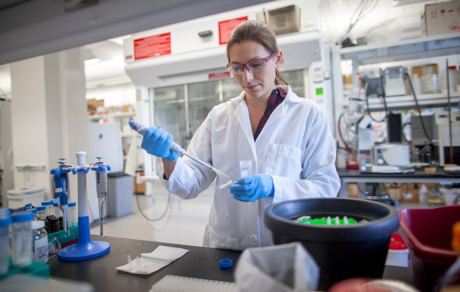 A researcher in a lab coat and glasses pipettes at lab bench