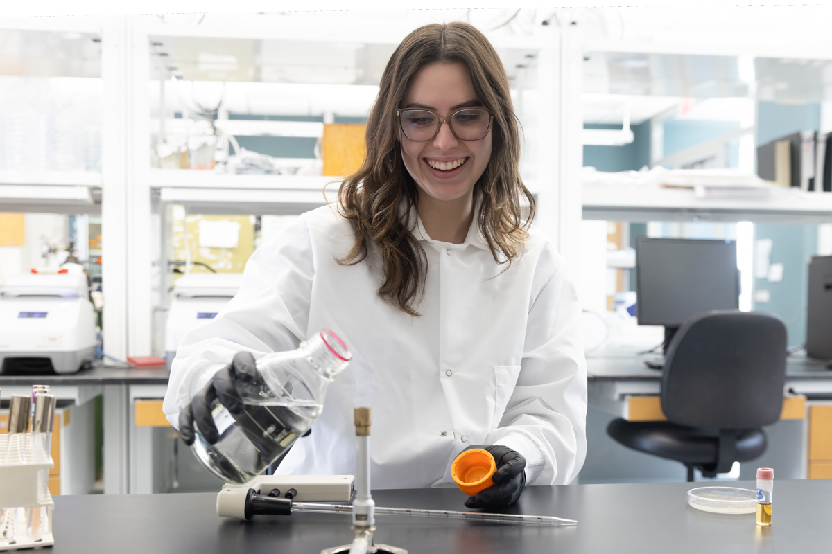 A woman in a white lab coat and latex gloves holds a flask of clear liquid in a laboratory