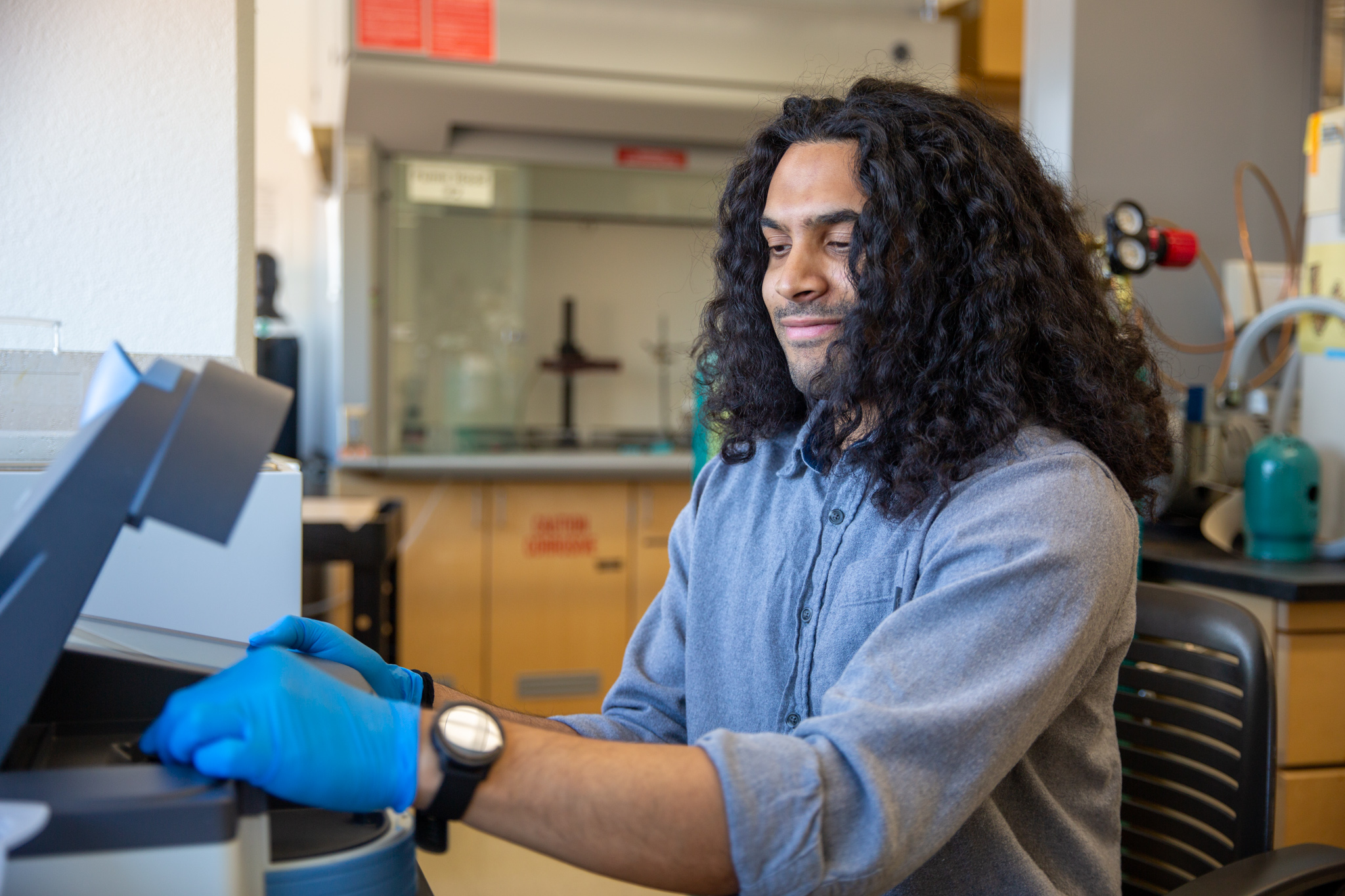 Daven is seated working on a lab bench, handling machinery with gloves.