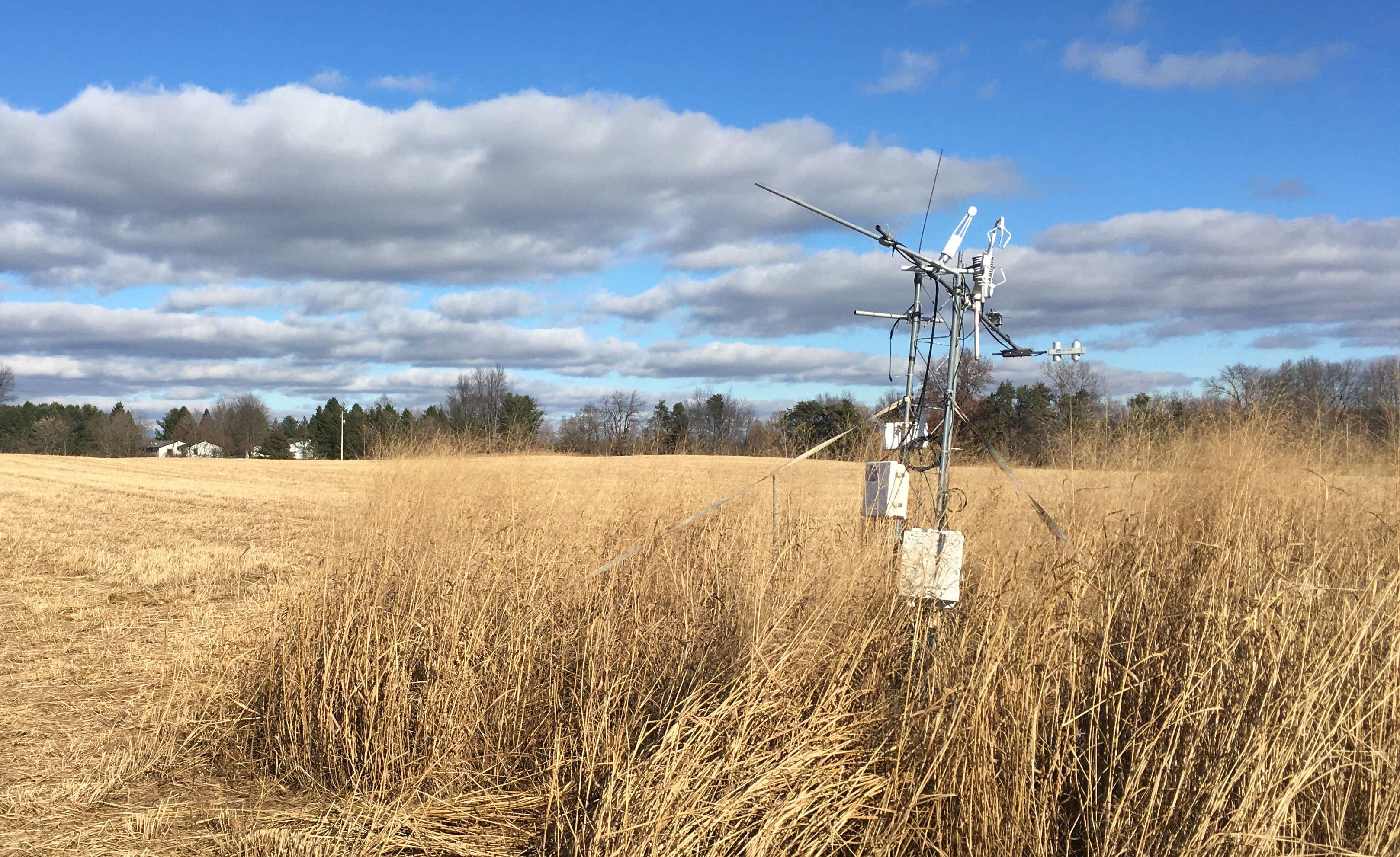 A tower with sensors and equipment mounted on it stands in a field of dried tan-colored vegetation, with trees visible in the background and a cloudy blue sky above.