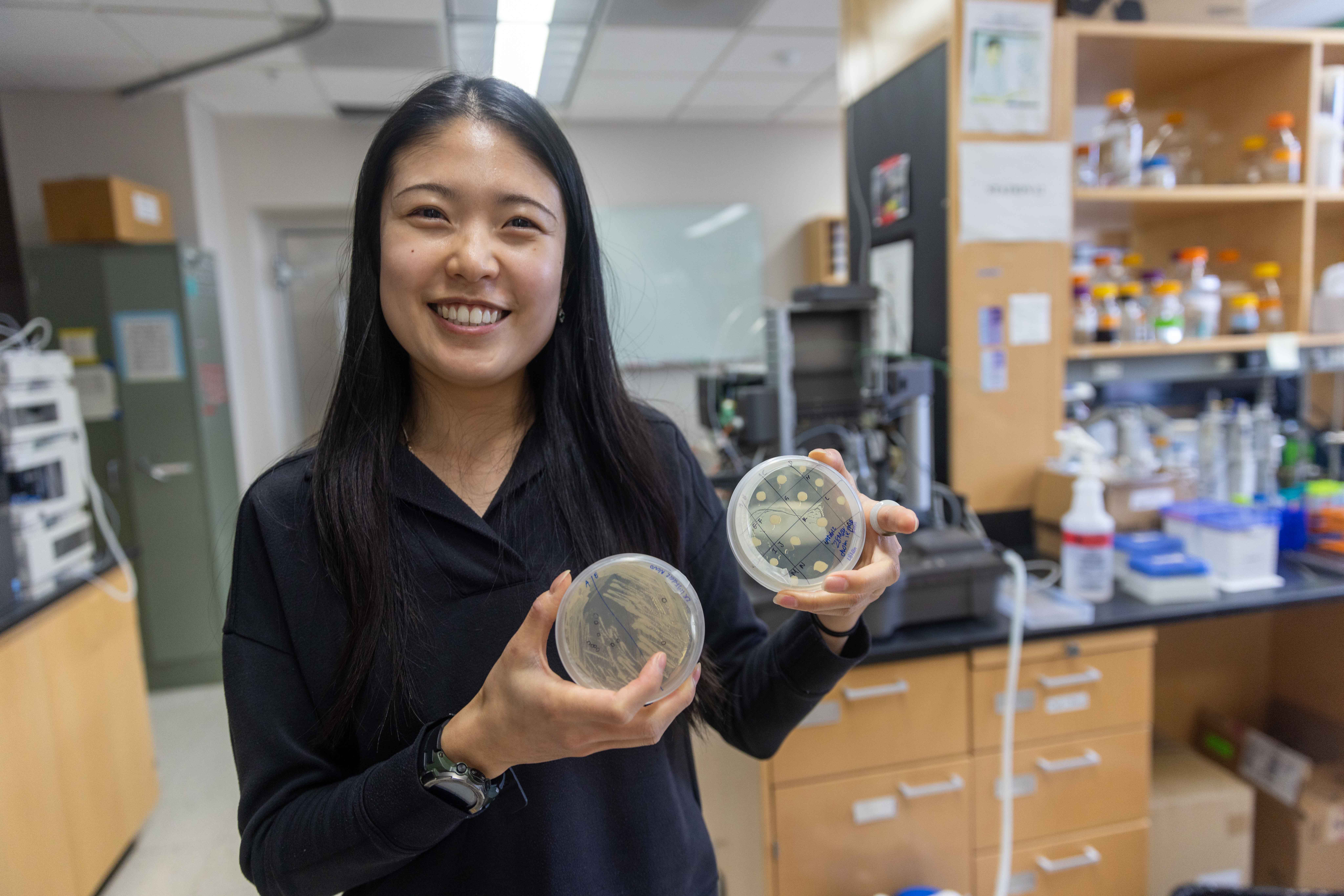 woman with long black hair standing in a lab holding two petri dishes