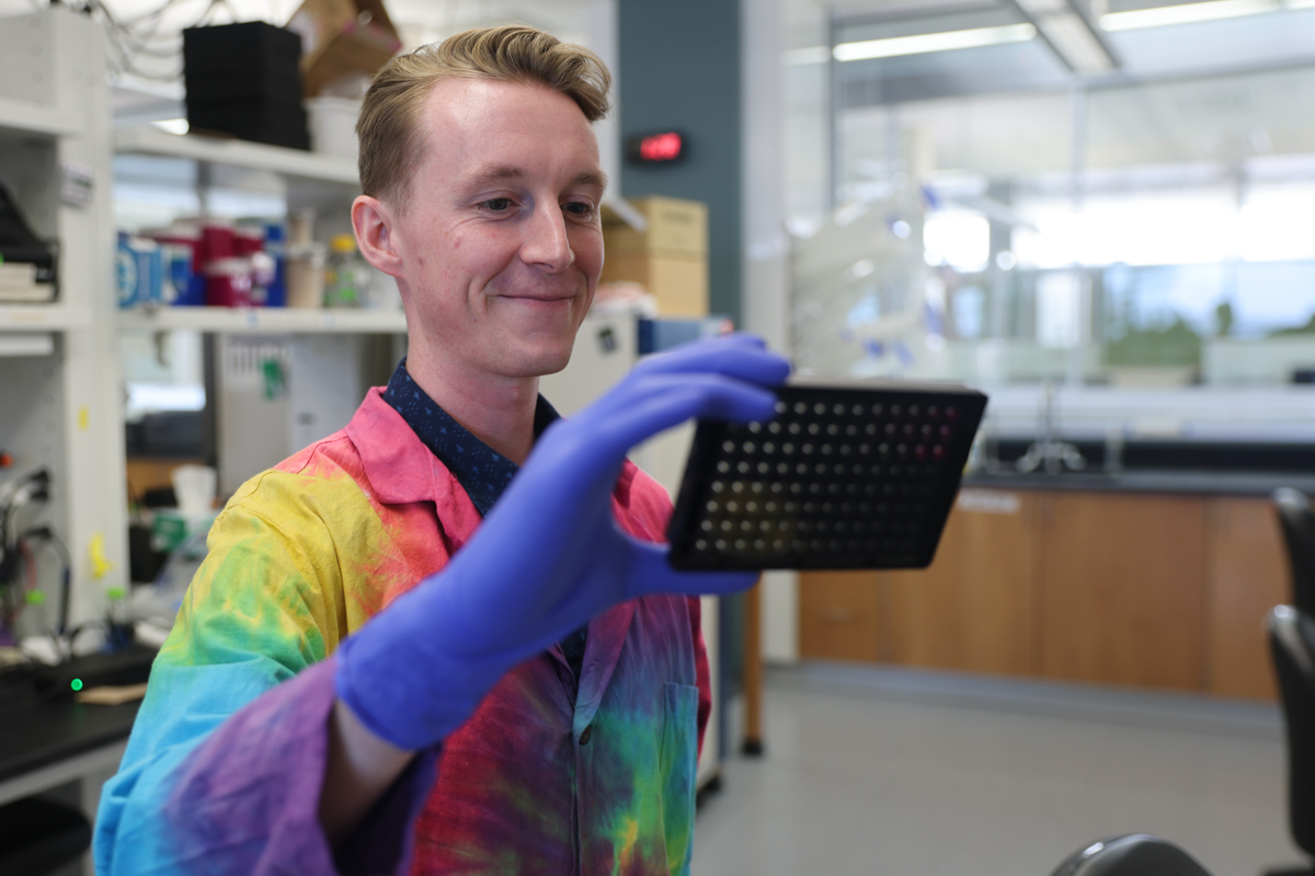 A blonde man in a tye-dyed lab coat holds a square tray in one blue gloved hand