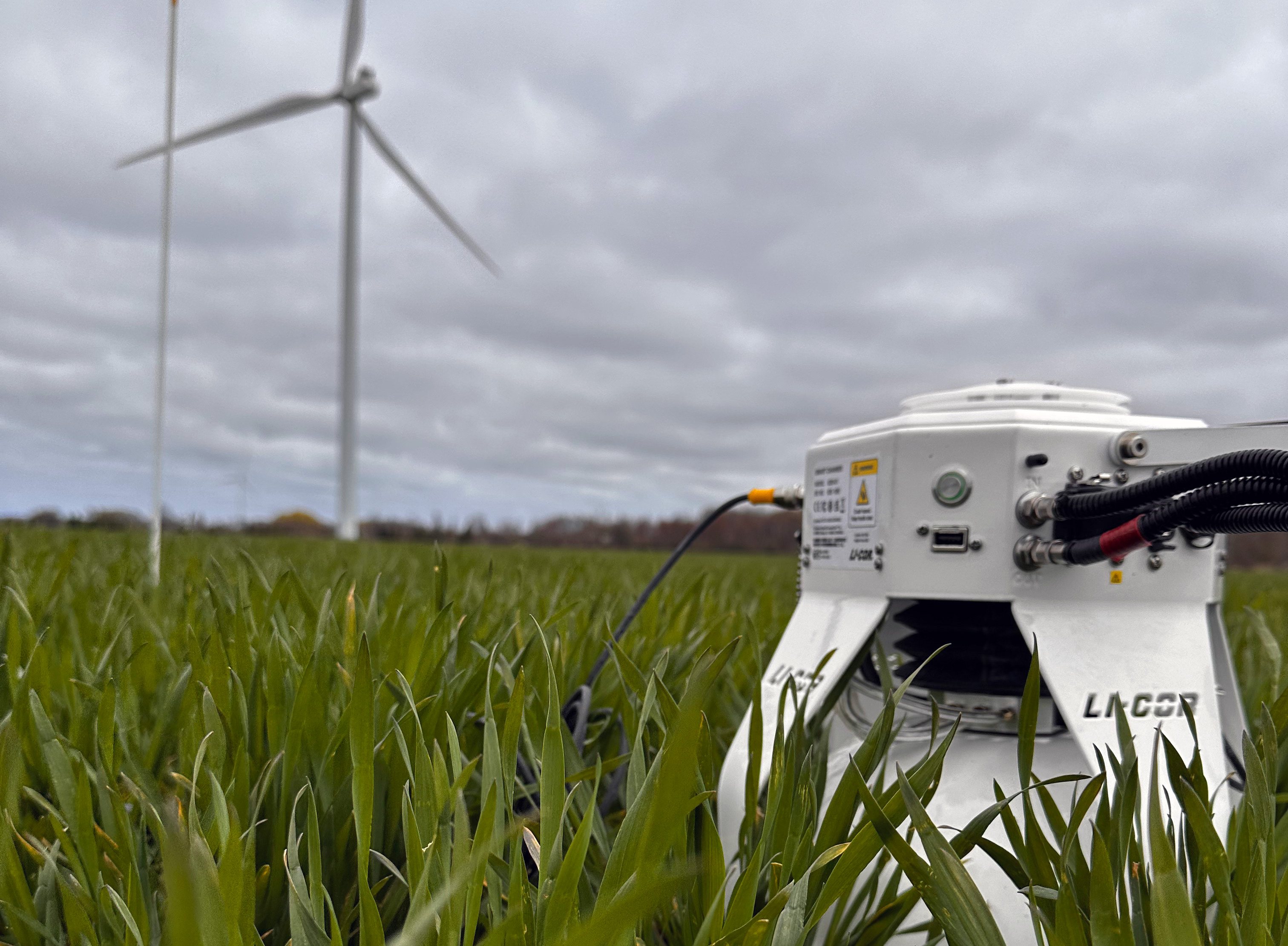 A LI-COR gas analyzer deployed at canopy level in a green crop field, with wind turbines visible in the background under an overcast sky.