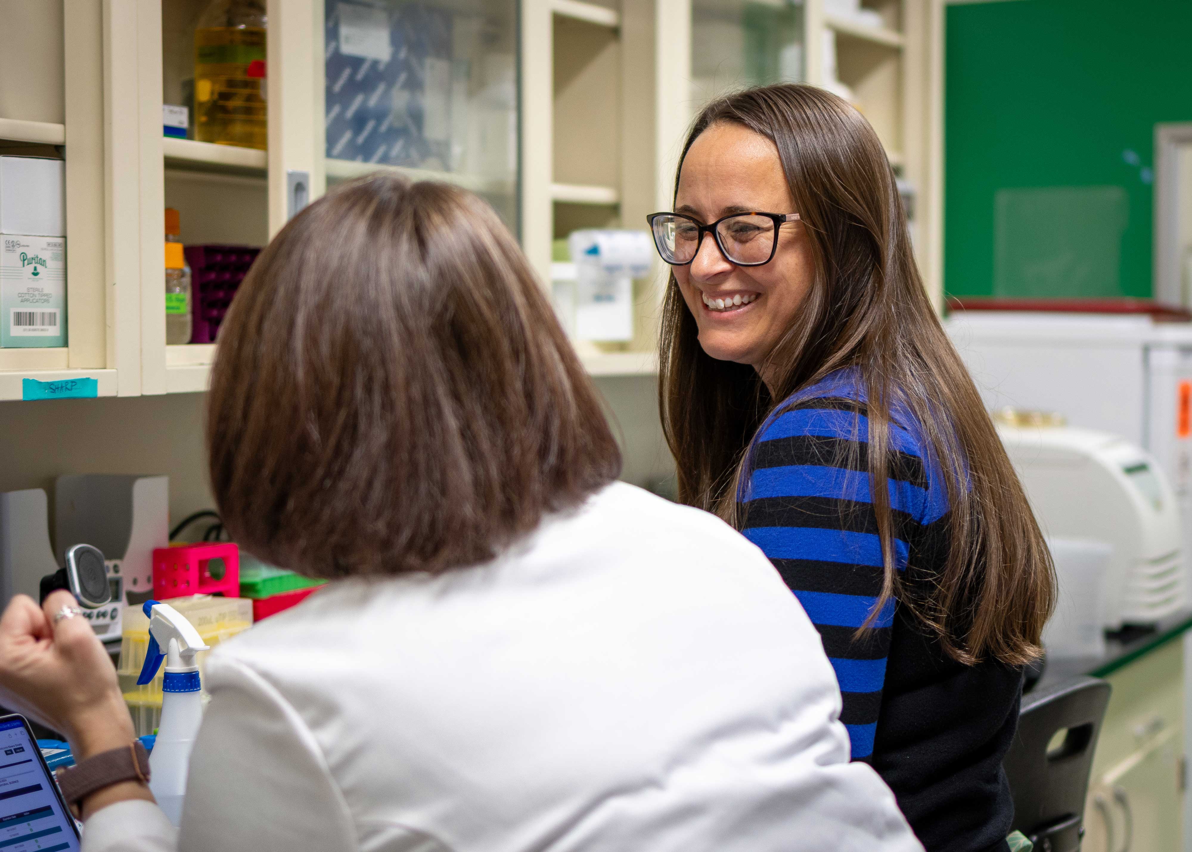 Two women seated at a lab bench. The one on the left is wearing a white lab coat and facing away from the camera. The other wears a blue shirt and glasses and is looking at the other.