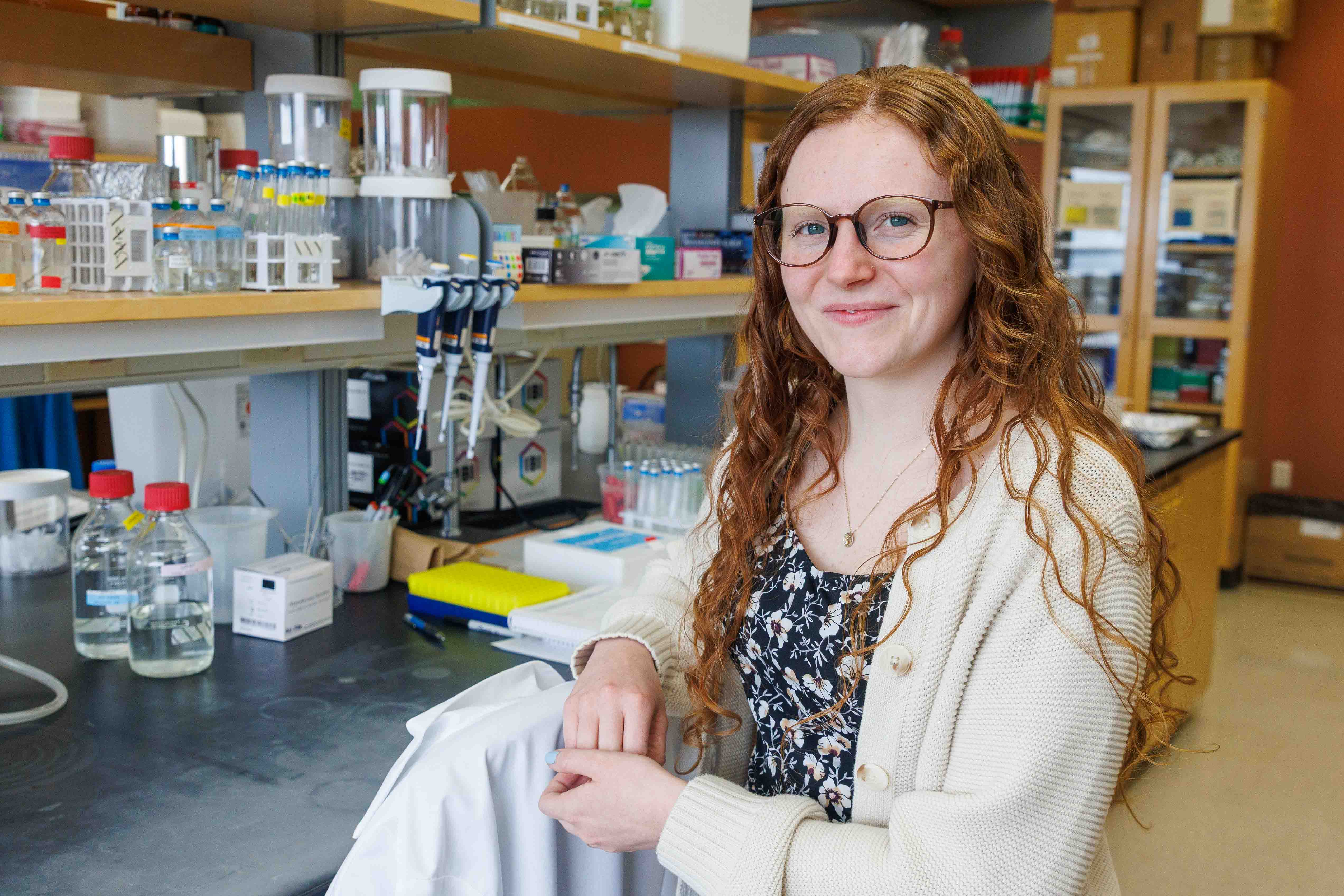 A researcher with long curly red hair and glasses, wearing a floral top and cream cardigan, smiles at the camera while seated in a lab surrounded by bottles, pipettes, and equipment.