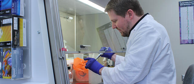 A man in a white lab coat and blue latex gloves pipettes liquid under a ventilation hood.