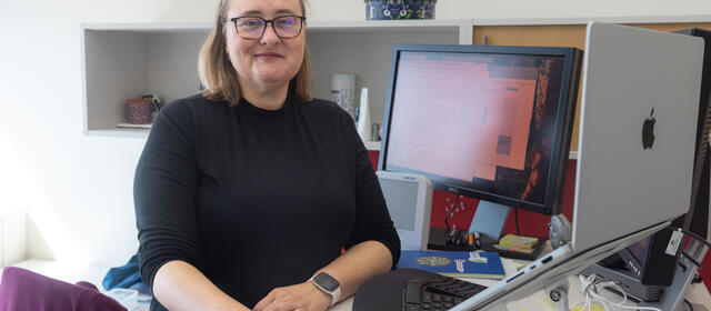 Woman with blonde hair and glasses sits at a desk with a computer monitor. She is turned to her right to face the camera