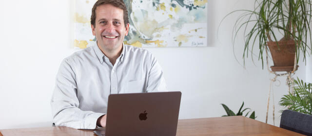 A smiling man sitting at a desk behind an open laptop computer