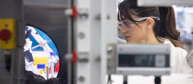 A researcher in a white lab coat and safety goggles looks through a circular portal into an automated laboratory workstation filled with robotic equipment and chemical vials.