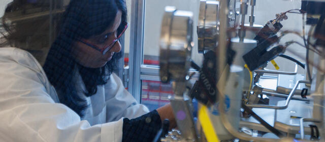 A researcher in a lab coat, safety glasses, and nitrile gloves sets up a high-pressure reactor system in a glass enclosure.