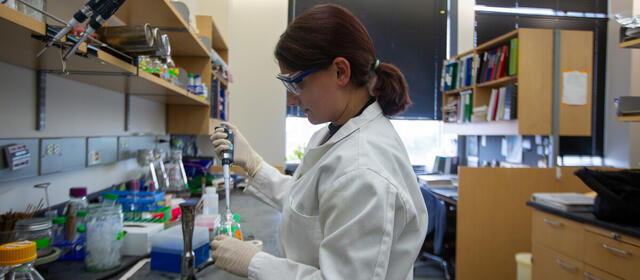A woman in a lab coat and safety glasses uses a pipette at a lab bench cluttered with jars, tubes, and supplies, with bookshelves visible behind her.
