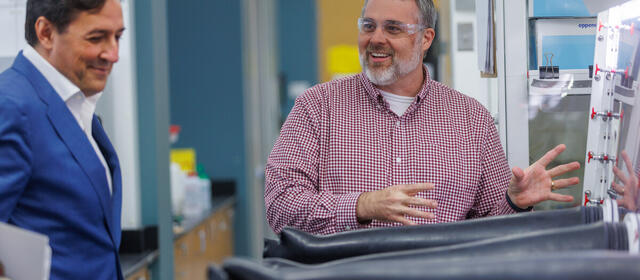 A man in a blue blazer looks on with a smile as a bearded man in a red checked shirt and safety glasses gestures animatedly beside a glovebox with black gloves in a laboratory.