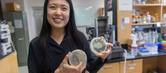 woman with long black hair standing in a lab holding two petri dishes