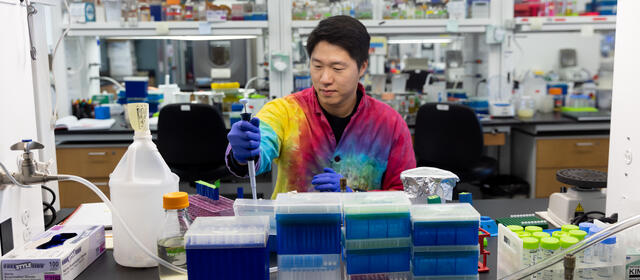 Asian man in tie-died lab coat seated at a lab bench