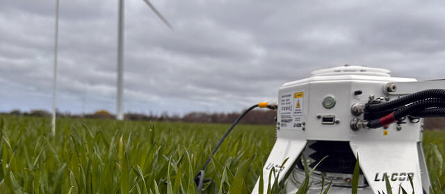 A LI-COR gas analyzer deployed at canopy level in a green crop field, with wind turbines visible in the background under an overcast sky.