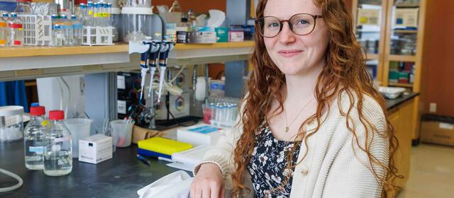 A researcher with long curly red hair and glasses, wearing a floral top and cream cardigan, smiles at the camera while seated in a lab surrounded by bottles, pipettes, and equipment.