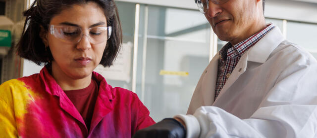 Two researchers wearing safety glasses examine colony plates, with one pointing to colony growth on a gridded agar plate held by the other.