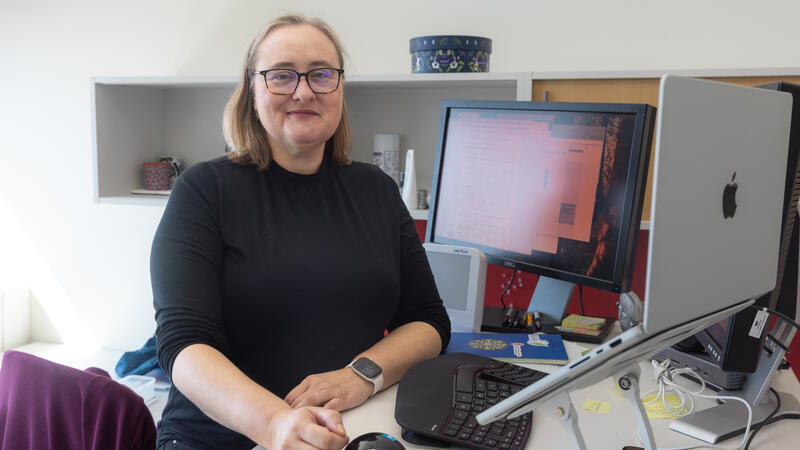 Woman with blonde hair and glasses sits at a desk with a computer monitor. She is turned to her right to face the camera