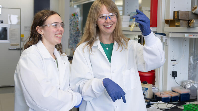 A woman in a white lab coat and safety glasses holds up a flask of opaque yellow liquid as another woman in similar attire looks on smiling