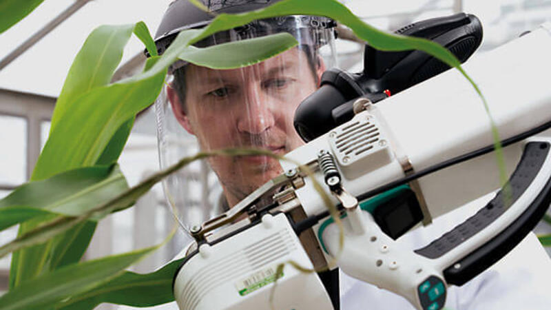 Man with a clear face visor looks at a piece of equipment. His face is framed by the instrument and green plant leaves