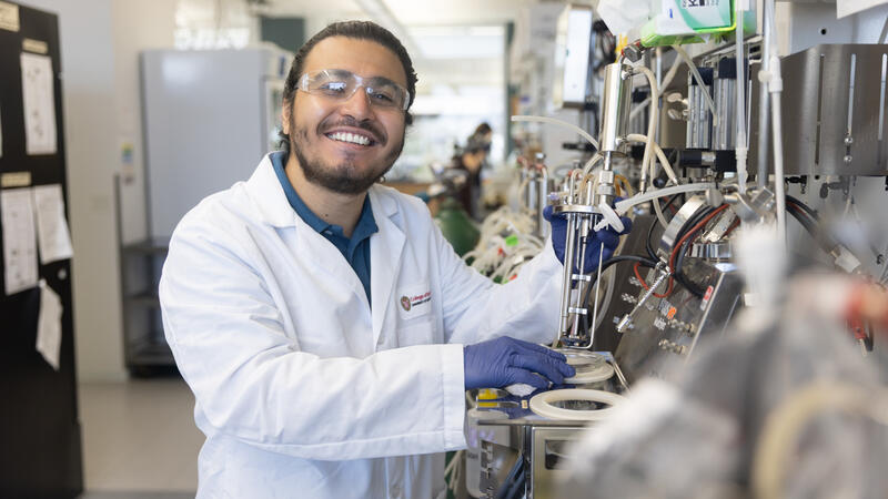 Smiling man in white lab coat and purple nitrile gloves holds a piece of equipment in a modern lab