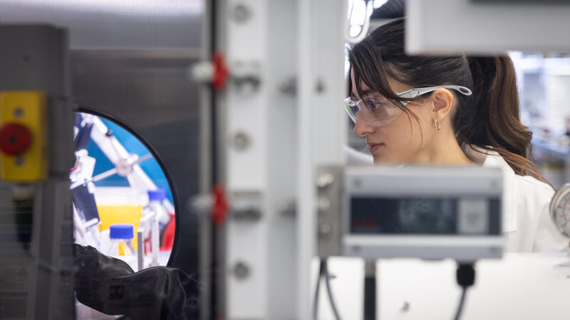 A researcher in a white lab coat and safety goggles looks through a circular portal into an automated laboratory workstation filled with robotic equipment and chemical vials.