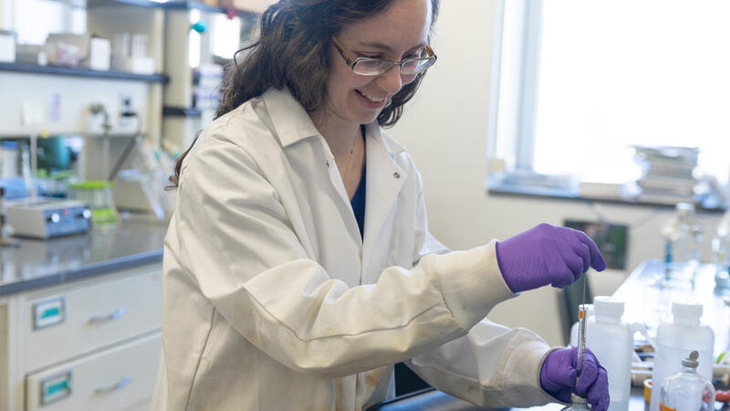 Woman in white lab coat and purple latex gloves opens a glass container on a black counter in a 