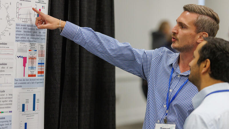 Man in blue and white shirt points to text on a poster as another man looks on