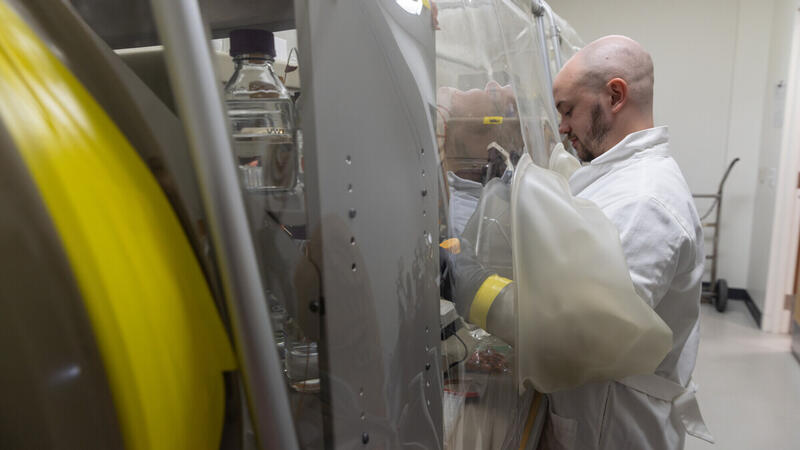 A man in a white lab coat stands in front of a clear plastic barrier with his arms extended through black rubber gloves that 
