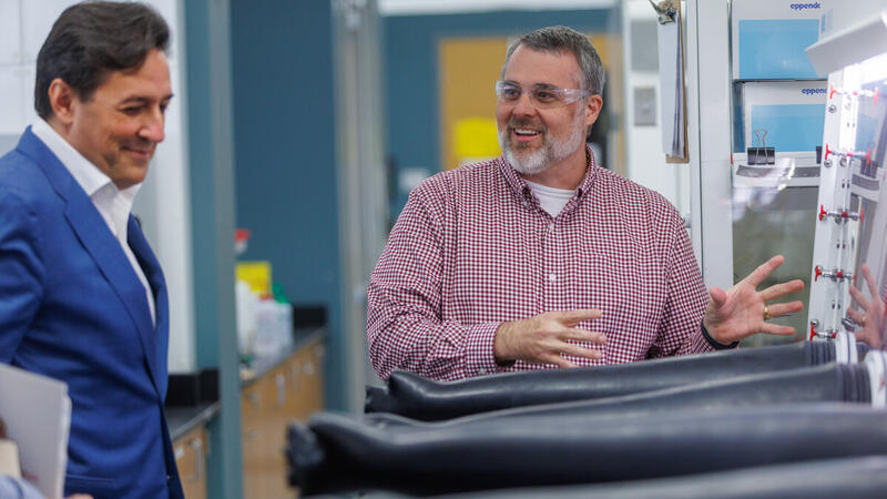 A man in a blue blazer looks on with a smile as a bearded man in a red checked shirt and safety glasses gestures animatedly beside a glovebox with black gloves in a laboratory.