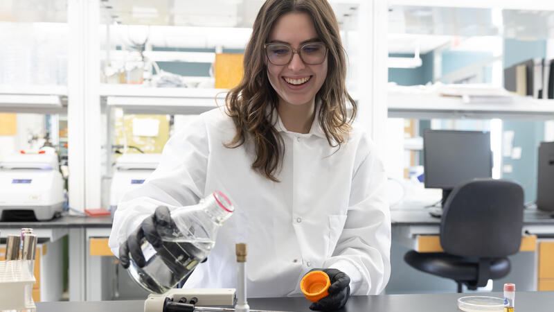 A woman in a white lab coat and latex gloves holds a flask of clear liquid in a laboratory