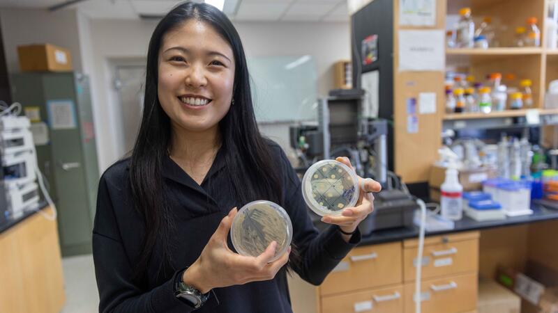 woman with long black hair standing in a lab holding two petri dishes