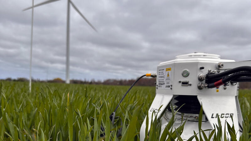 A LI-COR gas analyzer deployed at canopy level in a green crop field, with wind turbines visible in the background under an overcast sky.
