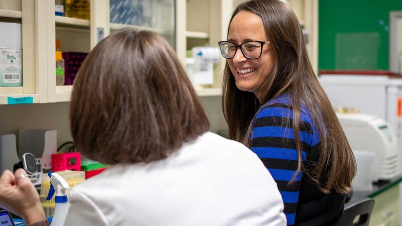 Two women seated at a lab bench. The one on the left is wearing a white lab coat and facing away from the camera. The other wears a blue shirt and glasses and is looking at the other.