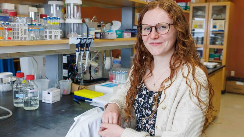 A researcher with long curly red hair and glasses, wearing a floral top and cream cardigan, smiles at the camera while seated in a lab surrounded by bottles, pipettes, and equipment.