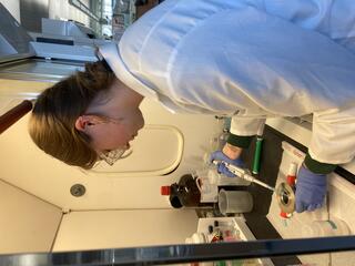 A woman in a lab coat and safety glasses turns away from the camera, holding a pipette and a metal container