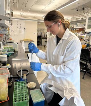 A woman in a lab coat and googles stands at a lab counter with a pipette and tube.